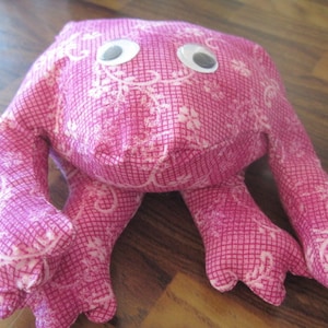 May include: A handmade pink plush toy frog with white floral patterns and googly eyes. The frog has four long limbs and a square head. The fabric has a grid pattern. The toy is designed to be soft and cuddly.