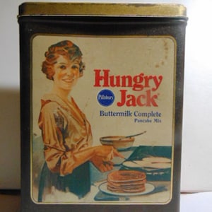 May include: Vintage Hungry Jack Buttermilk Complete Pancake Mix tin. The label features an illustration of a woman making pancakes. The tin is dark brown with a gold-colored top. Net weight is 32 oz (2 lb) or 907g.
