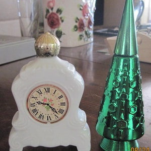 May include: Two vintage perfume bottles. One is white and shaped like a clock with Roman numerals. The other is green and shaped like a Christmas tree. Both bottles have a metallic finish.