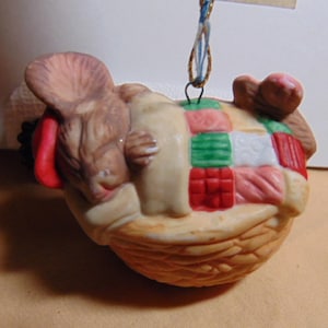 May include: A ceramic ornament featuring a sleeping mouse nestled in a woven basket. The mouse is light brown with a red accent, and rests on a patchwork quilt with red, green, and pink squares. A blue and gold string is attached for hanging.