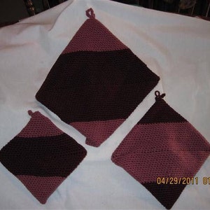 May include: Three crocheted pot holders in a dark brown and pink color scheme. The pot holders are square shaped with a loop at the top for hanging.