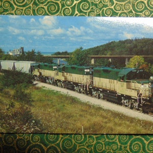 May include: A vintage postcard featuring a green and beige train with multiple cars traveling through a rural landscape. The train is traveling over a bridge and through a wooded area.