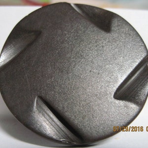 May include: A dark gray metal button with a textured surface and a raised, geometric design. The button has a slightly rounded shape.