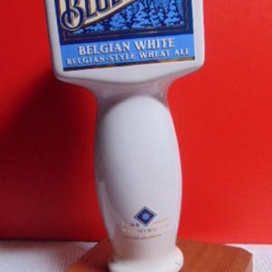 May include: A white and blue Blue Moon beer tap handle with the brand name and logo. The handle has a rectangular top with the words "Belgian White Belgian-Style Wheat Ale". It is mounted on a wooden base.
