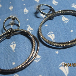 May include: A pair of silver hoop earrings with a double circle design. The earrings are made of metal and have a shiny finish.