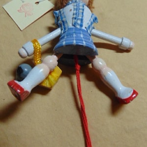 May include: A wooden pull-string doll with a painted face, wearing a blue and white checkered dress and red shoes. The doll has brown hair and a yellow beaded bracelet. A tag with a floral stamp hangs from the doll.
