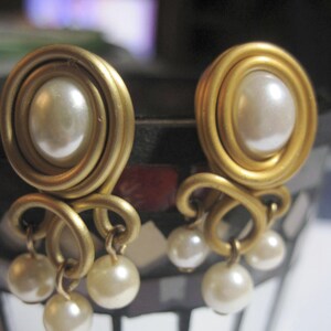 May include: A pair of gold-toned earrings featuring a central round faux pearl surrounded by a coiled wire frame. Below the main circular setting, three smaller faux pearls hang from individual gold-toned wire loops, creating a dangling effect.