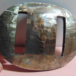 May include: Oval-shaped, iridescent buckle with two rectangular cutouts. The buckle has a mosaic-like appearance, with a cracked, textured surface in shades of brown, gray, and cream. The object is held by a person's fingers.