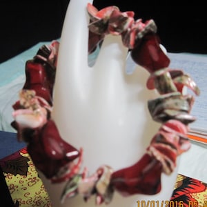 May include: A red and white bracelet made of small, flat, irregular pieces of material. The bracelet is on a white display stand.