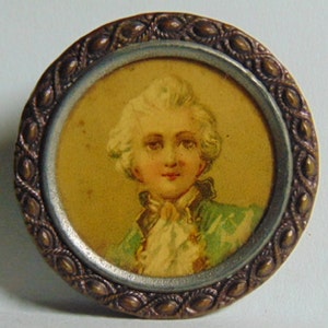 May include: A round, gold-colored metal button with a raised, ornate border. The center of the button features a portrait of a young person with white hair, wearing a green and white jacket.