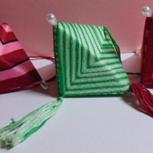 May include: Three handmade, colorful, geometric ornaments. The ornaments are square-shaped with a tassel at the bottom and a pearl bead at the top. One is red and pink, one is green and white, and the other is red and pink.