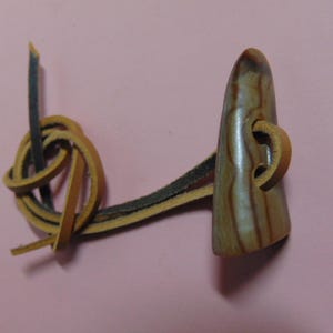May include: A wooden toggle button with a light brown, striped appearance. The button is connected to a leather loop and a knotted leather cord. The background is a solid pink color.
