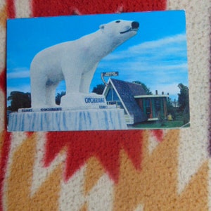 May include: A white polar bear statue stands on a platform with the text "Giant Cochrane" and "Cochrane, Ont.". The statue is in front of a building with a sign that reads "Cochrane Inn".