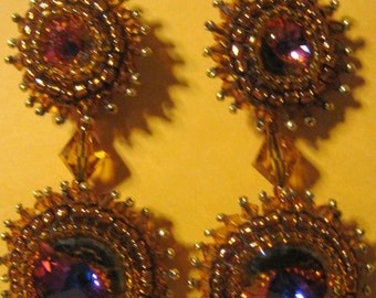May include: A pair of dangle earrings with two layers of beaded circles. The top layer is a smaller circle with a pink gem in the center. The bottom layer is a larger circle with a purple gem in the center. The earrings are made with gold-colored beads and have a gold-colored hook.