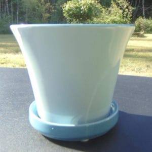May include: A light blue ceramic planter with a matching saucer. The planter has a flared shape, wider at the top and tapering towards the base. The saucer is round and slightly raised, designed to catch excess water. A decorative item for plants.