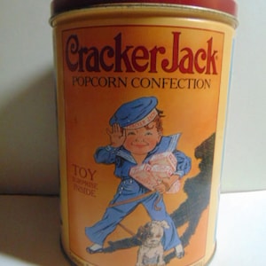 May include: Vintage Cracker Jack popcorn tin with a red lid and a yellow and orange label. The label features a cartoon boy in a sailor suit, a dog, and the text "Cracker Jack POPCORN CONFECTION". The tin contains 11oz of popcorn.
