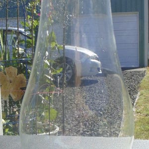 May include: A clear glass lamp chimney with a flared shape. The chimney has a wide base and tapers upwards. A blue label with the text "6016-5492" is visible near the base. The background shows a house and a car.