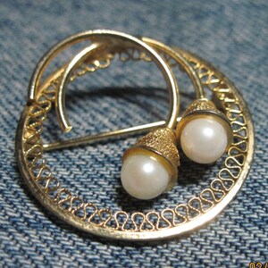 May include: A gold-toned brooch with a circular design featuring a delicate filigree pattern. Two white pearl-like stones are attached to the brooch.