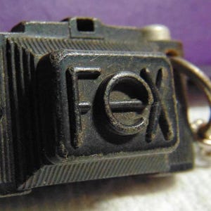 May include: A black metal camera keychain with the word "FOX" embossed on the front. The keychain has a silver top and a silver chain.