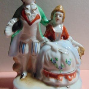 May include: A porcelain figurine depicting a couple in 18th-century attire. The man wears a green coat, white vest, and breeches with blue stripes. The woman is in a white dress with floral accents and a red shawl. The base is white with gold trim.