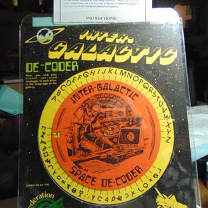 May include: A vintage "Inter-Galactic De-Coder" game with a circular dial featuring an astronaut illustration. The cover is black with yellow and orange accents, and includes the text "Space De-Coder" and "Federation of Planets."