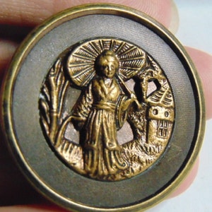 May include: A gold-toned metal button with a raised design of a person in a robe holding an umbrella, standing in front of a house. The button has a dark brown background.