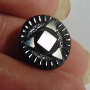 May include: Black glass button with a silver geometric design. The button has a raised, circular rim with a repeating pattern of small lines.