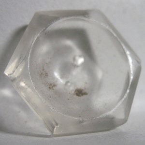 May include: A clear, hexagonal glass bottle stopper with a slightly rounded top. The stopper has a small, circular opening in the center.
