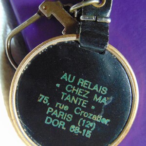 May include: A black leather keychain with a gold-colored metal ring and a round tag. The tag has the text "AU RELAIS • CHEZ MA TANTE. 75, rue Crozatier. PARIS (12°) DOR. 68-15" printed in green on a black background.