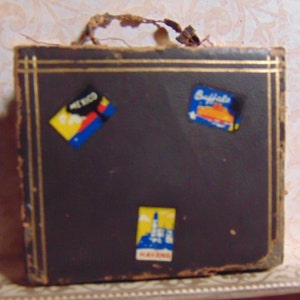 May include: A vintage brown suitcase with gold trim and three colorful travel stickers. The stickers say "Mexico", "Buffalo", and "Marlboro".