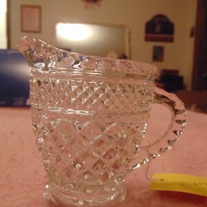 May include: A clear, vintage glass creamer with a diamond pattern. The creamer has a spout, a handle, and a round base. A yellow tag is attached to the handle. The creamer is likely from the mid-20th century.