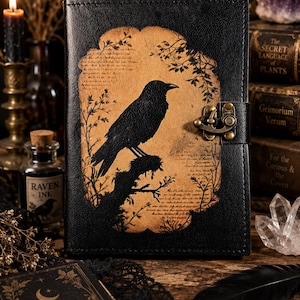 May include: A black leather-bound journal featuring a central tan-colored panel with a silhouette of a crow perched on a branch. The cover includes decorative botanical illustrations, faint script text, and a brass-colored metal latch closure.