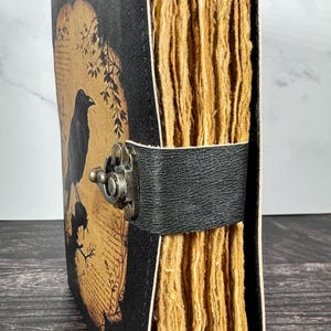 May include: A black journal with a textured cover featuring a graphic of a crow perched on a branch against a tan background. The journal has thick, deckled-edge tan pages and is secured by a black leather strap with a bronze-toned metal latch.