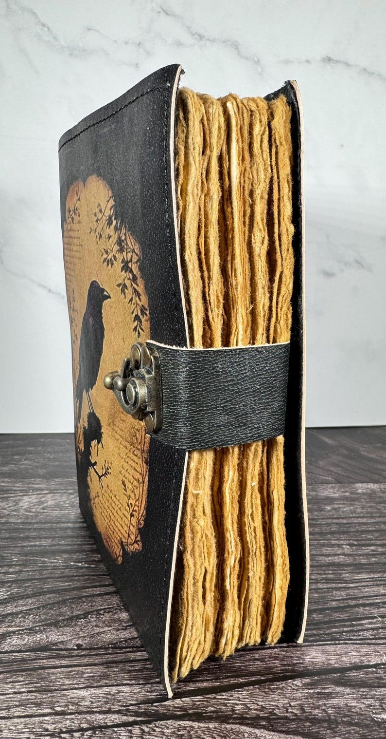 May include: A black journal with a textured cover featuring a graphic of a crow perched on a branch against a tan background. The journal has thick, deckled-edge tan pages and is secured by a black leather strap with a bronze-toned metal latch.
