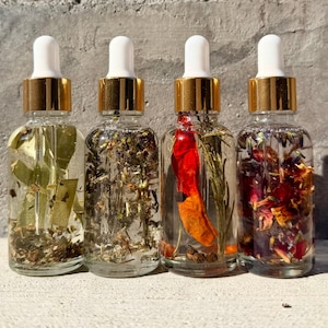 May include: Four clear glass dropper bottles with gold collars and white caps, each filled with a distinct liquid infusion. The bottles contain various botanical elements including green leaves, dried herbs, a red chili pepper, and colorful flower petals.
