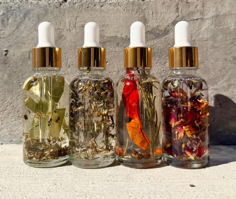 May include: Four clear glass dropper bottles with gold collars and white caps, each filled with a distinct liquid infusion. The bottles contain various botanical elements including green leaves, dried herbs, a red chili pepper, and colorful flower petals.