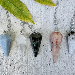 May include: Five faceted gemstone pendulums hanging from silver-toned metal chains. The stones include a matte light blue, a clear translucent crystal, a dark black stone with green flecks, a speckled peach-colored stone, and a white stone.