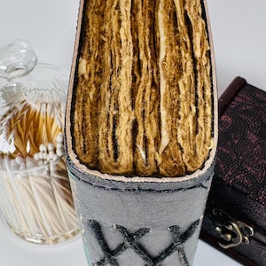 May include: A handmade journal with a gray leather cover and stitched detailing. The pages are thick and have a textured, aged appearance. The journal is partially open, revealing the rough-cut edges of the pages. A glass jar with matches is in the background.
