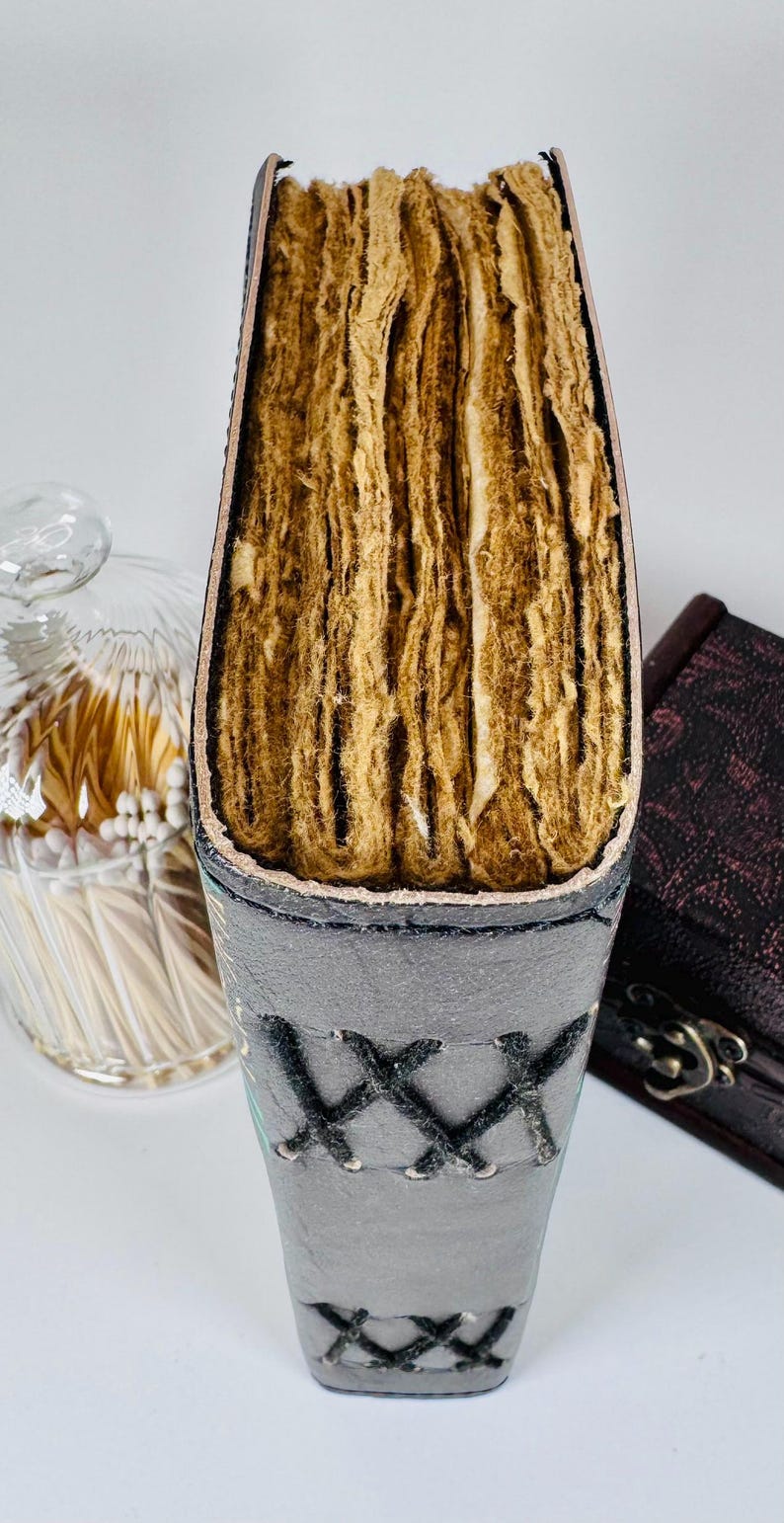 May include: A handmade journal with a gray leather cover and stitched detailing. The pages are thick and have a textured, aged appearance. The journal is partially open, revealing the rough-cut edges of the pages. A glass jar with matches is in the background.