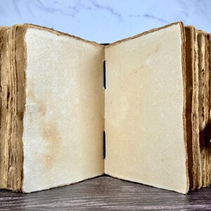 May include: An open journal with thick, cream-colored, deckle-edged paper pages. The book features a dark cover and a metal latch closure on the side. The binding is visible in the center, showing black stitching holding the pages together.