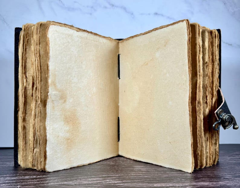 May include: An open journal with thick, cream-colored, deckle-edged paper pages. The book features a dark cover and a metal latch closure on the side. The binding is visible in the center, showing black stitching holding the pages together.