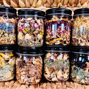 Puede incluir: Ocho frascos de vidrio llenos de hierbas y productos botánicos secos, cada uno con una tapa negra. Los frascos contienen una variedad de ingredientes coloridos, incluyendo elementos azules, rosas, amarillos y naranjas. Los frascos están dispuestos sobre una superficie tejida.