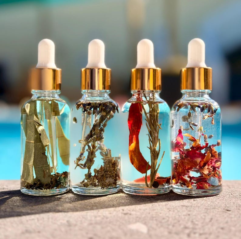 May include: Four clear glass dropper bottles with gold collars and white rubber bulbs, each filled with a clear liquid and different botanical elements including green leaves, dried lavender, a red chili pepper, and dried flower petals.
