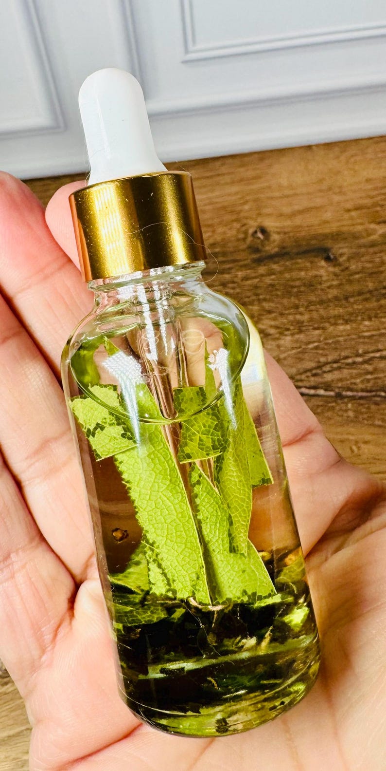 May include: A clear glass dropper bottle filled with a light yellow liquid and several green leaves submerged inside. The bottle features a gold-colored metal collar and a white rubber dropper top, providing a functional design for dispensing the liquid contents.
