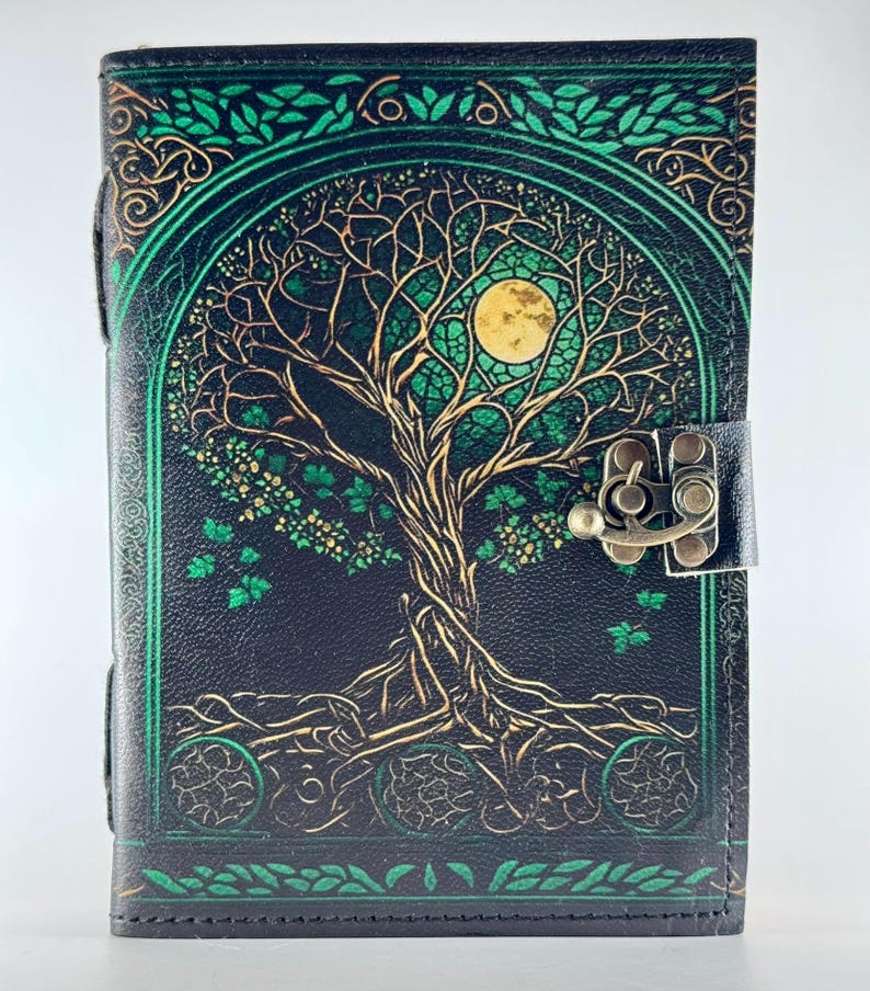 May include: A black leather journal with a tree of life design. The cover features a detailed illustration of a tree with a full moon, in shades of gold and green. The journal has a clasp closure.