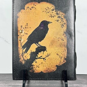 May include: A black leather journal cover featuring a central rectangular graphic of a crow perched on a branch against a warm, golden-yellow background. The design includes faint, illegible script text and two small brass-colored metal rivets on the left.