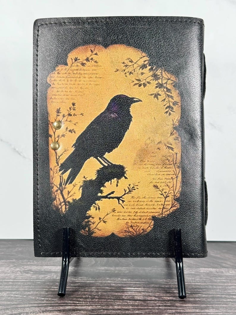 May include: A black leather journal cover featuring a central rectangular graphic of a crow perched on a branch against a warm, golden-yellow background. The design includes faint, illegible script text and two small brass-colored metal rivets on the left.