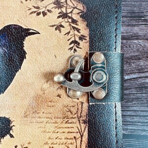 May include: A journal with a black textured spine and a tan cover featuring a dark raven illustration, botanical branch silhouettes, and script text. The cover is secured by a metallic antique brass-toned latch with a swivel locking mechanism.
