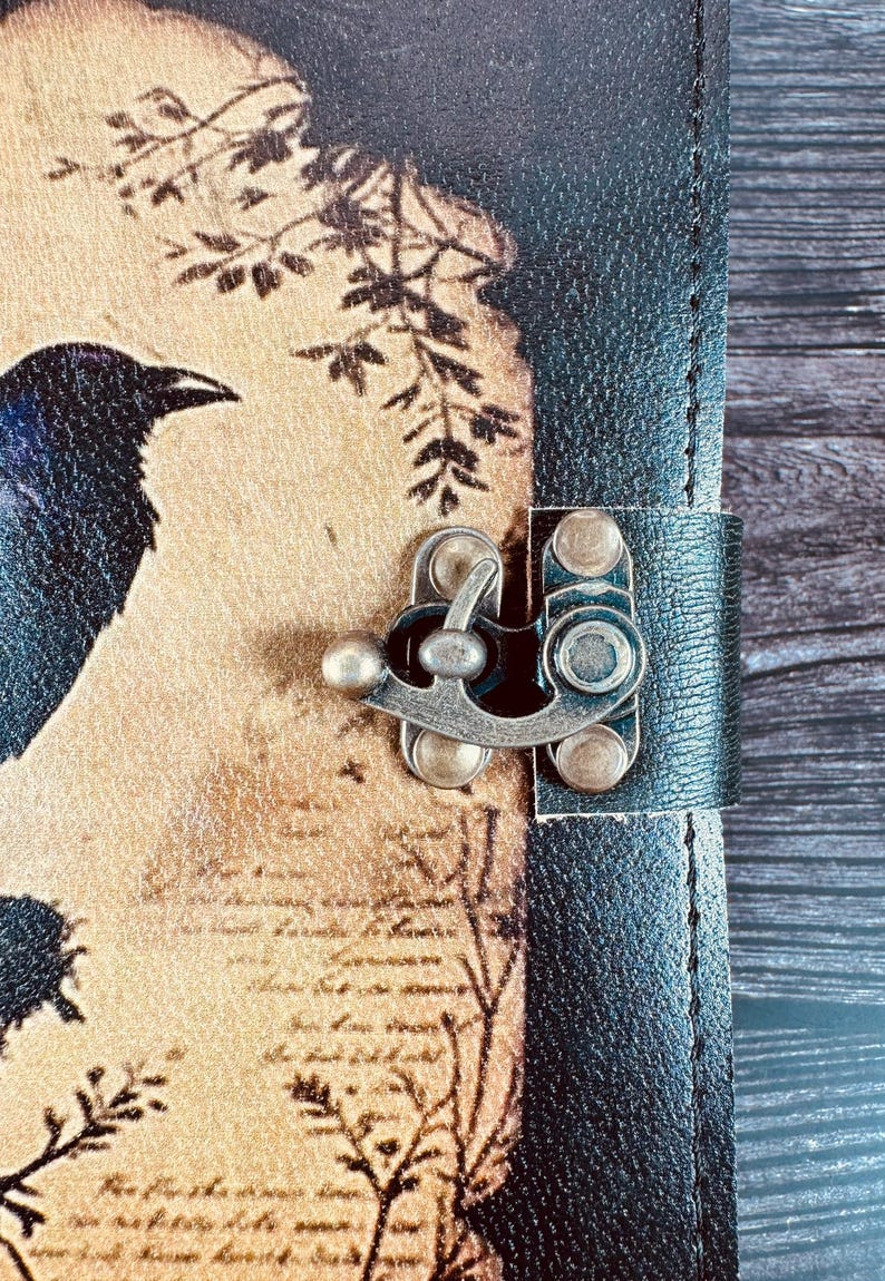 May include: A journal with a black textured spine and a tan cover featuring a dark raven illustration, botanical branch silhouettes, and script text. The cover is secured by a metallic antique brass-toned latch with a swivel locking mechanism.