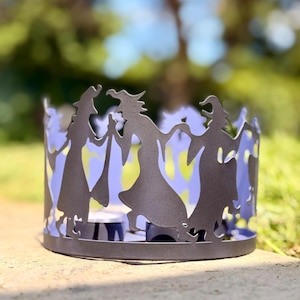 May include: A cylindrical metal candle sleeve featuring a cut-out silhouette design of multiple witches holding hands in a circle. The exterior surface is finished in a matte dark gray color, while the interior shows a light purple hue.